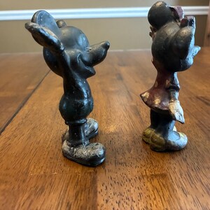 Op de afbeelding: Twee vintage gietijzeren beeldjes van Mickey en Minnie Mouse. Mickey staat in een klassieke pose met uitgestrekte armen. Minnie draagt een jurk met een strik. Beide figuren zijn geschilderd in zwart, rood en wit.