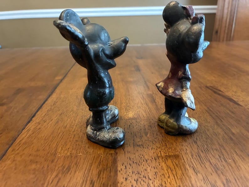 Op de afbeelding: Twee vintage gietijzeren beeldjes van Mickey en Minnie Mouse. Mickey staat in een klassieke pose met uitgestrekte armen. Minnie draagt een jurk met een strik. Beide figuren zijn geschilderd in zwart, rood en wit.