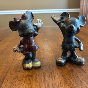 Op de afbeelding: Twee vintage gietijzeren beeldjes van Mickey en Minnie Mouse. Minnie draagt een rode jurk met witte stippen en een strik. Mickey draagt een korte broek. Beide figuren hebben uitgestrekte armen en zijn donker geschilderd.