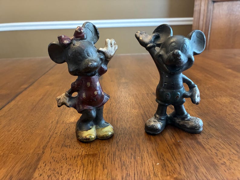 Op de afbeelding: Twee vintage gietijzeren beeldjes van Mickey en Minnie Mouse. Minnie draagt een rode jurk met witte stippen en een strik. Mickey draagt een korte broek. Beide figuren hebben uitgestrekte armen en zijn donker geschilderd.