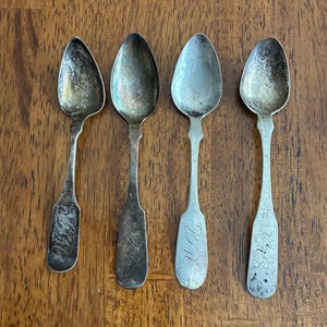 May include: Four antique silver spoons with oval bowls and ornate handles are arranged on a wooden surface. The spoons show signs of age and wear, with tarnishing and engraved details. The spoons are a vintage kitchenware item.