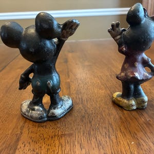 Op de afbeelding: Twee vintage gietijzeren beeldjes van Mickey en Minnie Mouse. Mickey staat in een klassieke pose met &eacute;&eacute;n arm omhoog. Minnie draagt een polkadot jurk. Beide figuren hebben een verweerde, verouderde uitstraling.