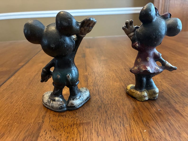Op de afbeelding: Twee vintage gietijzeren beeldjes van Mickey en Minnie Mouse. Mickey staat in een klassieke pose met &eacute;&eacute;n arm omhoog. Minnie draagt een polkadot jurk. Beide figuren hebben een verweerde, verouderde uitstraling.