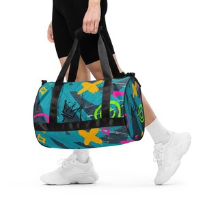 May include: A blue and black duffel bag with a colorful graffiti design. The bag has a black strap and a crown graphic.