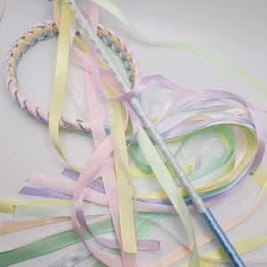 May include: A pastel fairy wand with a blue bow and a braided hoop. The wand is silver with a blue handle and adorned with pastel ribbons in shades of pink, yellow, green, and lavender. The hoop is braided with similar pastel colours.