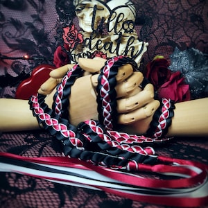 Puede incluir: Una cinta trenzada negra y roja, sostenida por una mano de madera, con el texto "Till Death" arriba. La cinta está entrelazada con acentos blancos y rojos. Rosas rojas y un objeto en forma de corazón en el fondo.