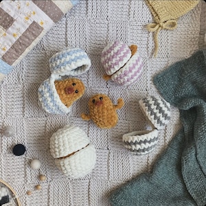 May include: A collection of six crocheted eggs, each with a different design. The eggs are in various shades of blue, white, and brown. The eggs are arranged on a white knitted surface.