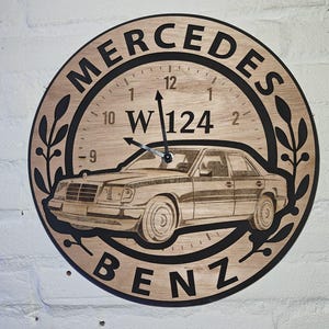 Può includere: Orologio da parete in legno con un design di auto Mercedes-Benz W124 inciso al laser. Il quadrante dell'orologio include le parole "MERCEDES BENZ" e "W 124" in un cerchio, con numeri e lancette nere.