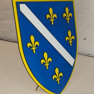 May include: A shield-shaped emblem featuring the flag of Bosnia and Herzegovina. The shield has a blue background with a white diagonal stripe and six yellow fleur-de-lis symbols. The border is yellow.