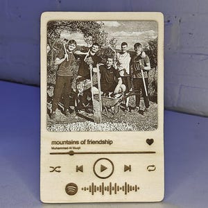 Puede incluir: Placa de madera con una imagen grabada con láser de seis personas, con el texto "mountains of friendship" y el nombre del artista. El diseño se asemeja a una interfaz de reproductor de música, con iconos de reproducción, salto y me gusta.