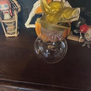 Könnte beinhalten: Eine klare Glasflasche mit Korken, gekrönt von einem goldfarbenen Schweinekopf. Die Flasche steht auf einer dunklen Holzoberfläche. Im Hintergrund befinden sich weitere dekorative Gegenstände, darunter eine Puppe in einem Korbstuhl.