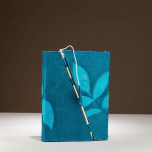 Op de afbeelding: Een turquoise notitieboek met een bladerenpatroon. Het notitieboek is gesloten met een diagonale houten stok en wit touw. De kaft heeft een textuur, en het bladerenontwerp is een lichtere tint turquoise. Ongeveer 15 cm hoog.