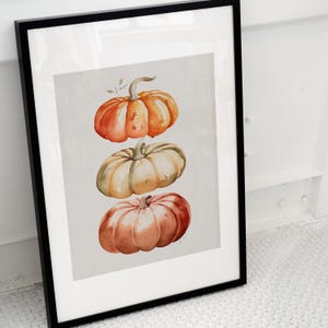 May include: A framed watercolour print featuring three stacked pumpkins in shades of orange, muted green, and reddish-brown. The artwork is set against a light grey background and enclosed within a simple, thin black frame with a white mat border.