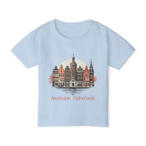 Może przedstawiać: Jasnoniebieska koszulka z kolorową ilustracją domów na kanałach Amsterdamu. Projekt zawiera różne style i kolory budynków, z tekstem "Amsterdam Netherlands" w kolorze różowym.