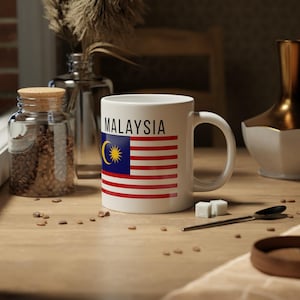 以下が含まれることがあります： マレーシアの国旗の上に「MALAYSIA」の文字が書かれた白いセラミックマグカップ。国旗のデザインは、赤と白のストライプ、三日月と星のある青いカントン、黄色の星で構成されています。マグカップは木製の表面に置かれています。