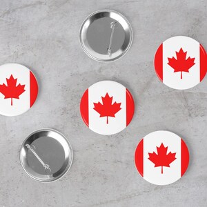 May include: Five round pins with a red maple leaf on a white background. The pins are arranged in a circle on a grey surface. One pin is silver and has a pin back.