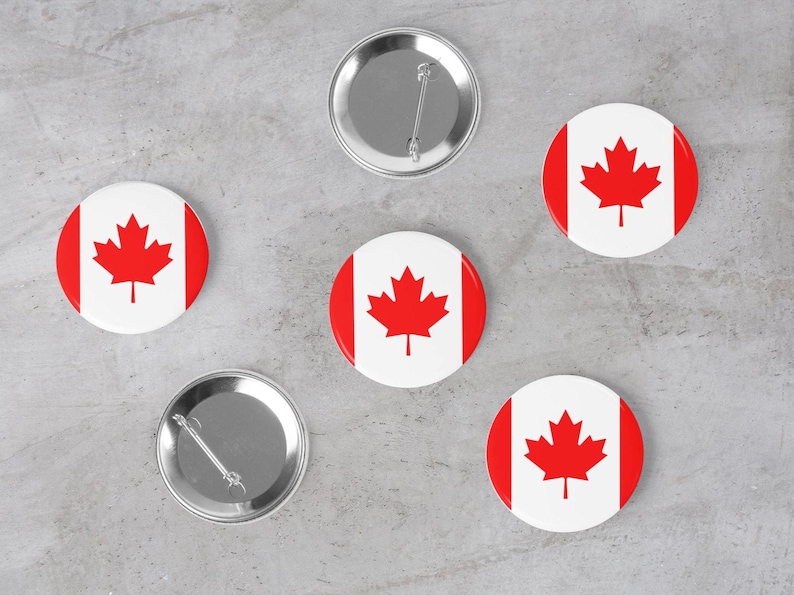 May include: Five round pins with a red maple leaf on a white background. The pins are arranged in a circle on a grey surface. One pin is silver and has a pin back.