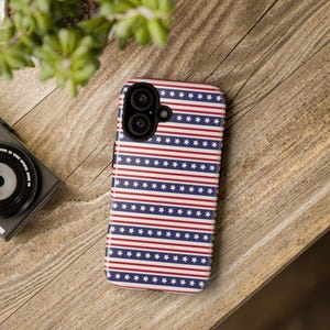 May include: A smartphone case with a patriotic design featuring alternating red and white stripes and rows of white stars on a navy blue background. The case is lying on a wooden surface.