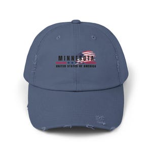 May include: A distressed blue baseball cap featuring the word "MINNESOTA" in black, with a stylized American flag graphic. Below the text is "UNITED STATES OF AMERICA" in black. The cap has a pre-curved bill and a worn appearance.