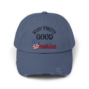 May include: A distressed blue baseball cap with the text "STAY PRETTI GOOD America" in black and red. The peace sign is colored with the American flag. The cap has a pre-curved visor and distressed details.