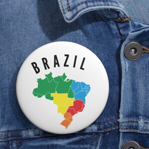 May include: A white button with the word "BRAZIL" in black, arched text. The button features a colorful map of Brazil, with regions in green, yellow, red, blue, and orange. The button is set against a denim background.
