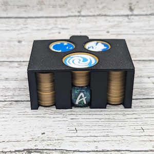 May include: A black plastic storage box with three compartments, each with a different colored circular design. The box is filled with gold-colored tokens and a blue and green dice with the letter 'A' on it.