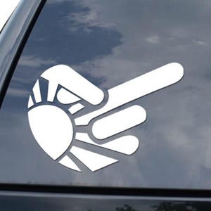 May include: White vinyl car decal featuring a stylized hand gesture with a sun design. The decal is applied to the rear window of a black vehicle. The design is a unique and eye-catching graphic.