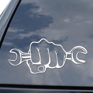 May include: White decal of a fist holding a wrench, applied to a black vehicle window. The design features a hand clenching a wrench, with the wrench's ends visible on either side of the fist. The decal is a simple, bold graphic.