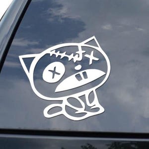 May include: White vinyl sticker featuring a cartoon cat with stitched head and cross-shaped eyes. The sticker is applied to the rear window of a black vehicle. The cat has a unique, slightly distressed design.