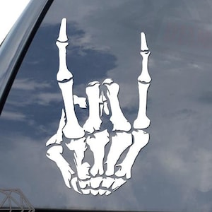 May include: White skeleton hand making the rock and roll hand gesture. The hand is on a car window.