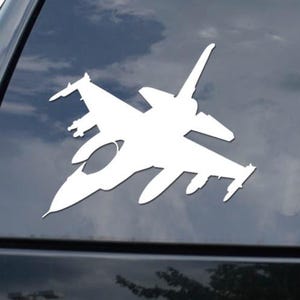 May include: White decal of a fighter jet on a black car window. The jet is a detailed silhouette, showcasing wings, tail, and fuselage. The background shows a cloudy sky. A diamond logo is visible.