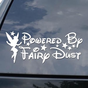 May include: White vinyl decal applied to a car window. The decal features a silhouette of a fairy, stars, and the words "Powered By Fairy Dust" in a whimsical font. The background shows a cloudy sky.