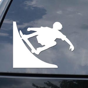 May include: White vinyl decal of a snowboarder performing a trick on a ramp. The snowboarder is in mid-air, with a helmet and athletic attire. The decal is applied to a vehicle's rear window, with a cloudy sky visible in the background.