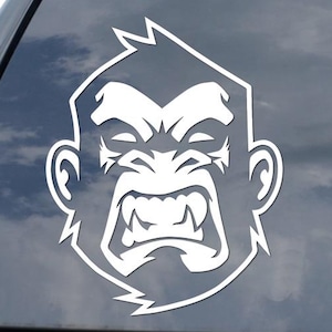 May include: White vinyl decal of a gorilla's face with an angry expression. The decal is applied to the rear window of a black vehicle. The gorilla's face is a bold, graphic design.