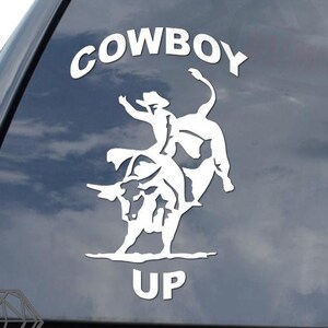 May include: White vinyl decal with a silhouette of a cowboy riding a bucking bull. The decal says "COWBOY UP" in a white, bold font.