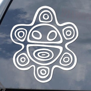 May include: White vinyl sticker featuring a stylized, symmetrical design resembling a sun or face. The design is composed of concentric circles and curved lines. The sticker is affixed to the rear window of a black vehicle.