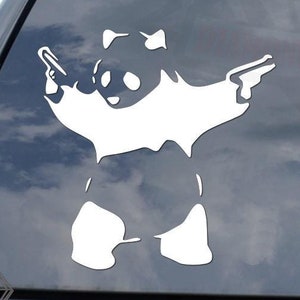 May include: A white vinyl sticker of a panda bear holding two guns. The sticker is on the back window of a black car.
