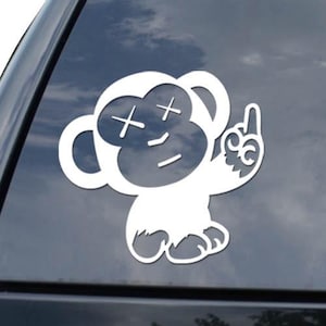 May include: White vinyl sticker of a cartoon monkey giving the middle finger. The monkey has crossed-out eyes and a neutral expression. The sticker is applied to the rear window of a black vehicle.