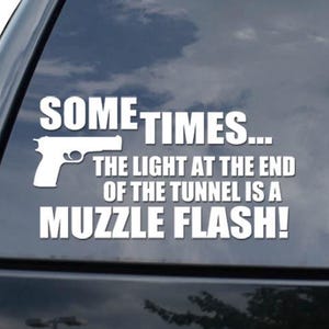 May include: White vinyl decal with the text "SOMETIMES... THE LIGHT AT THE END OF THE TUNNEL IS A MUZZLE FLASH!" and a graphic of a handgun. The decal is applied to the rear window of a black vehicle.