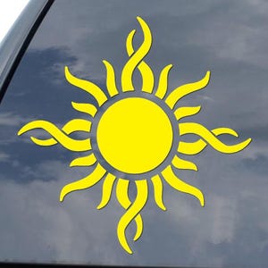 May include: A yellow sun-shaped decal with stylized rays and a central circle, affixed to a car window. The design features a bright yellow color, creating a cheerful and eye-catching aesthetic. The decal is likely for vehicle decoration.