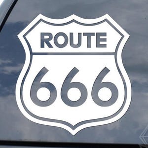 May include: White Route 666 sticker on a car window. The sticker features a shield design with the word "ROUTE" above the number "666". The sticker is a light gray color and is placed on a dark gray car window.