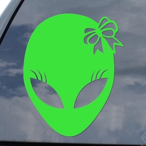 May include: A neon green alien head sticker with a bow on top. The alien has large, almond-shaped eyes and a smooth, oval head. The sticker is displayed on a car window.