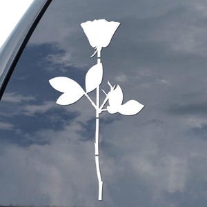 May include: A white rose decal, featuring a stylized flower with a long stem and leaves, is affixed to a car window. The design is a simple, elegant silhouette, suitable for car decoration. The background shows a cloudy sky.