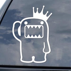 May include: White vinyl decal of a cartoon monster wearing a crown, waving with one arm, and showing its teeth. The decal is applied to a car window. The background is a cloudy sky.