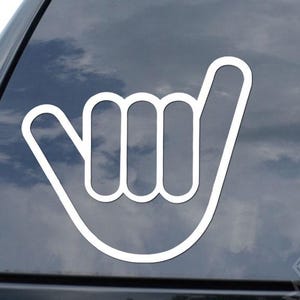 May include: White vinyl decal of the shaka hand gesture applied to a car window. The shaka symbol is a hand gesture often associated with surfing and the Aloha spirit. The decal is on a black car with a cloudy sky in the background.