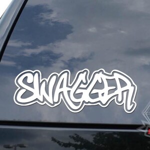 May include: White "SWAGGER" sticker in a graffiti style font, applied to the rear window of a black vehicle. A diamond logo is visible below the text. The sky is reflected in the window.
