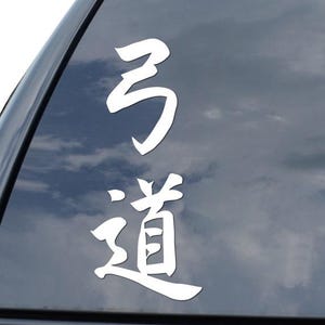 May include: White vinyl decal with Japanese characters, affixed to the rear window of a black vehicle. The characters are stylized and appear to read "Kyudo" or "The Way of the Bow". The decal is positioned on the upper left side of the window.