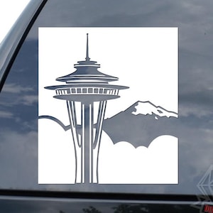 May include: A gray vinyl decal featuring the Space Needle and a mountain range. The Space Needle is a prominent landmark in Seattle, Washington. The decal is applied to a car window.