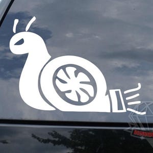 May include: White vinyl decal of a snail with a turbocharger shell. The snail has a stylized design with a turbine wheel and exhaust details. The decal is applied to a car window.