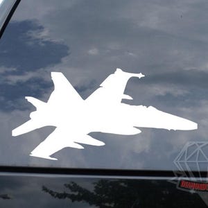 May include: A white decal of a fighter jet is affixed to the rear window of a black vehicle. The jet's silhouette is visible against a cloudy sky. The decal is a detailed representation of the aircraft.